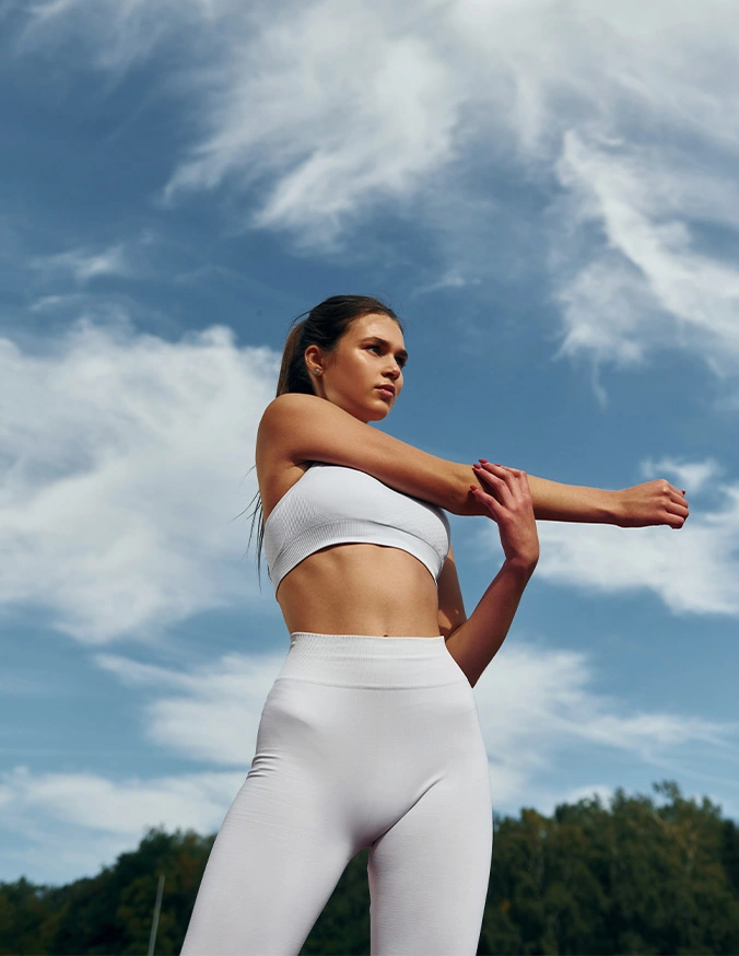 Woman in white athletic wear stretching outdoors against blue sky with clouds during workout - Awake lipo risks in Brookfield and Appleton, WI.