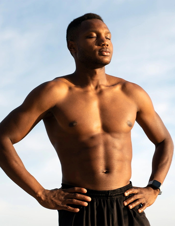 Fit shirtless man standing confidently outdoors with hands on hips against blue sky background - Male chest gynecomastia in Brookfield and Appleton, WI.