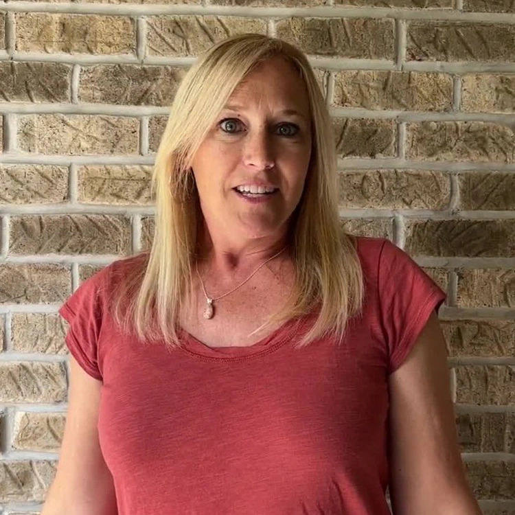 Woman with blonde hair in coral shirt standing against brick wall sharing patient testimonial