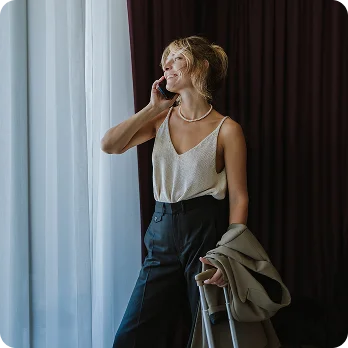 Woman in white tank top and black pants talking on phone while standing by window with curtains - in Brookfield and Appleton, WI.
