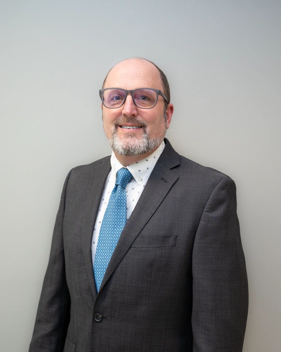 Dr. Conrardy in dark suit and blue tie standing against white wall for corporate portrait